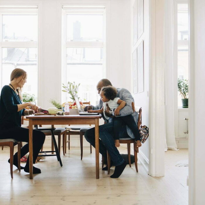 Äiti, isä ja taapero viettävät yhdessä vapaa aikaansa pöydän äärellä. Äiti tekee ruokaa ja isä selaa tablettia taapero sylissään Retta