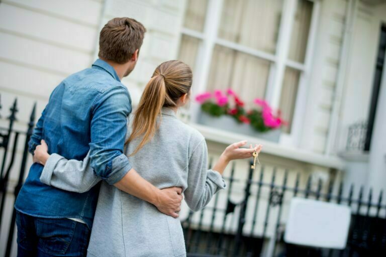 Nainen ja mies ovat uuden asuntonsa edessä avaimet kädessä kainaloittain Retta